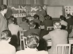  Assemblea, un militare parla a un gruppo di uomini in una sala adibita a scuola guida.